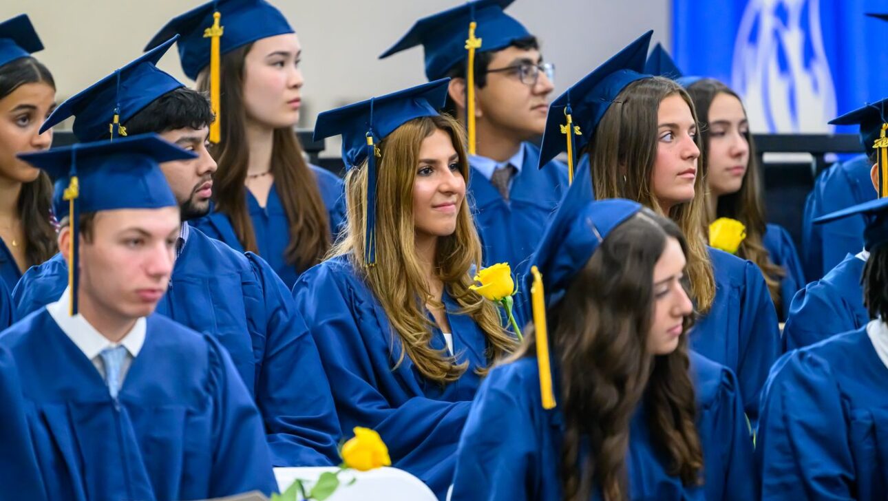 A group of graduates wearing blue caps and gowns sit in rows, some holding yellow roses, during a graduation ceremony.