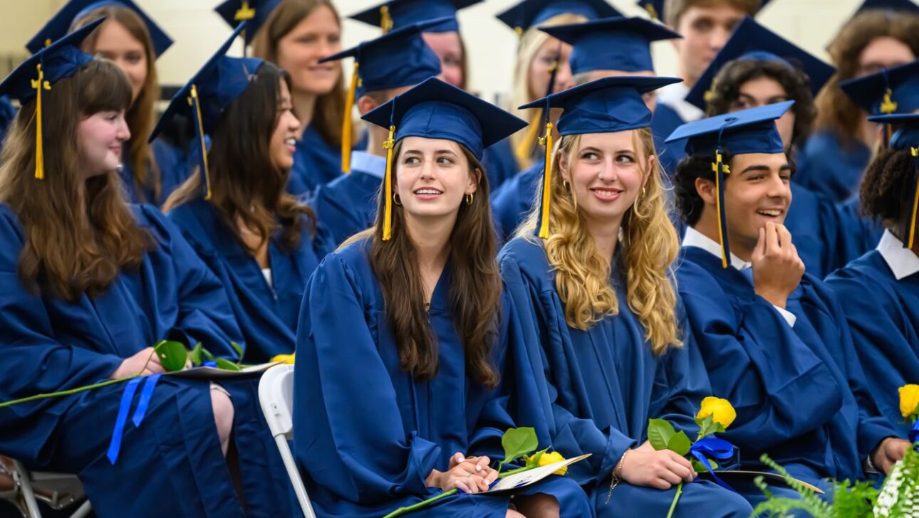 Students in blue graduation gowns and caps sit together, holding flowers, during a commencement ceremony.