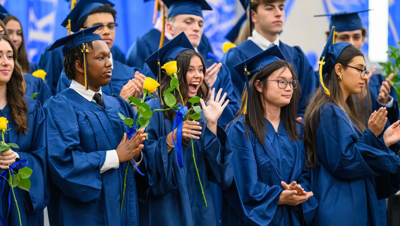 A group of students in blue graduation gowns hold yellow flowers, with some clapping and one student cheering enthusiastically in the middle.
