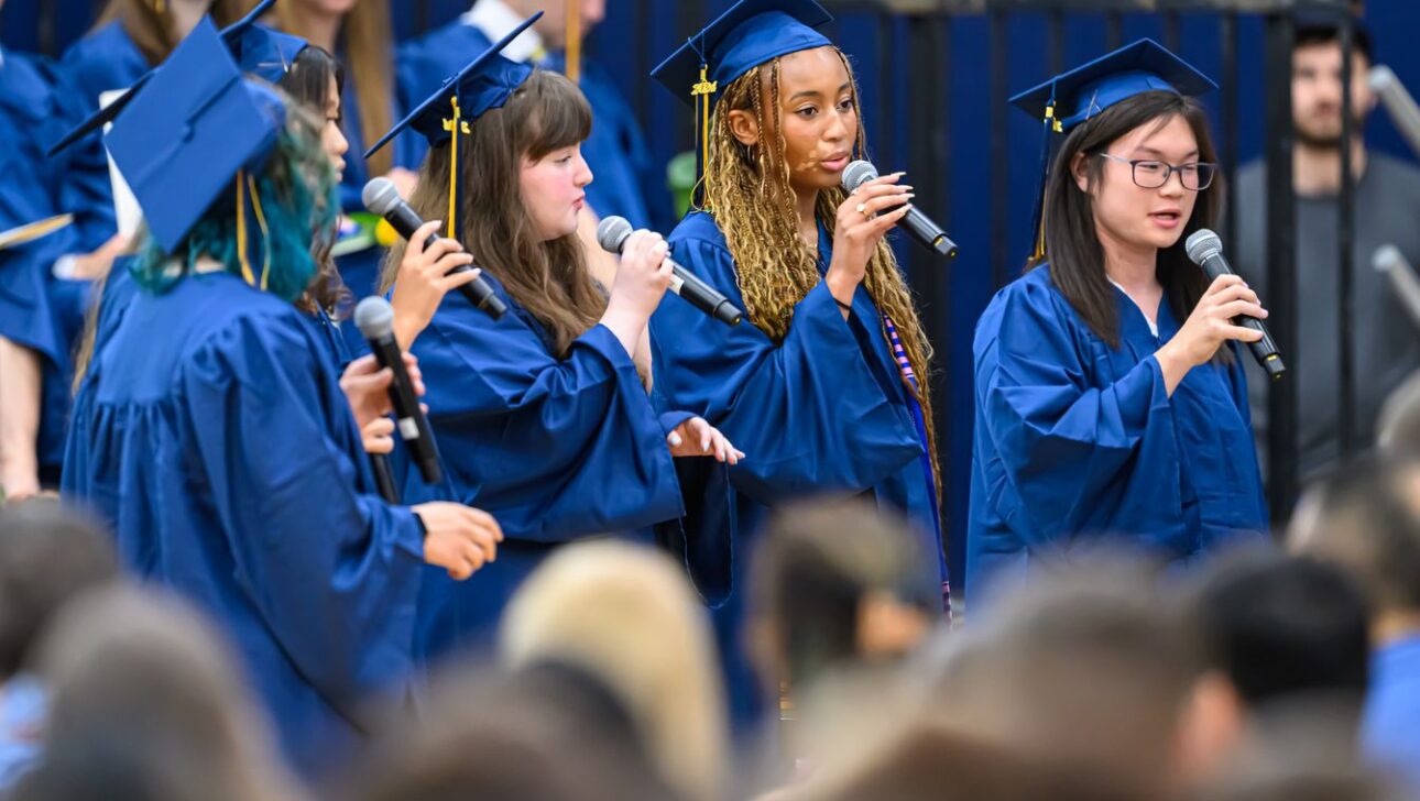 A group of people in blue graduation gowns and caps sing into microphones during a ceremony, with onlookers in the foreground.