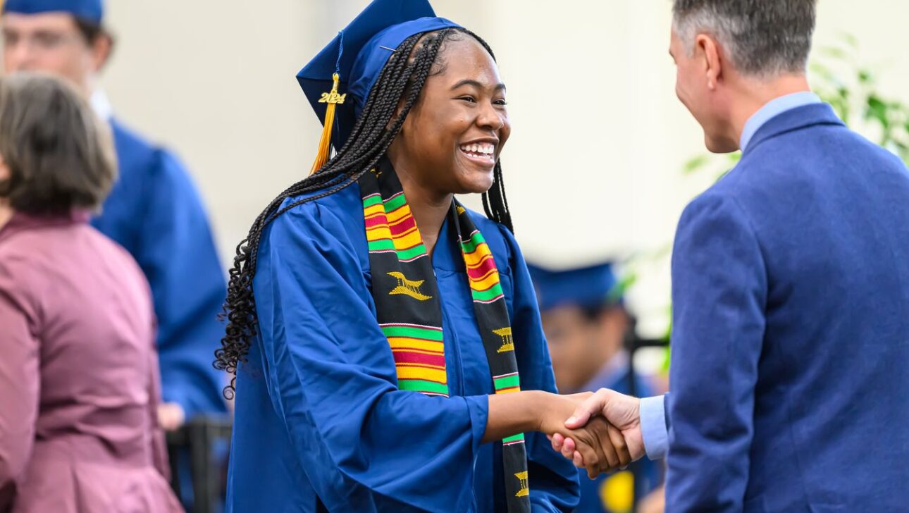 A graduate in blue cap and gown shakes hands and smiles with a man during a graduation ceremony.