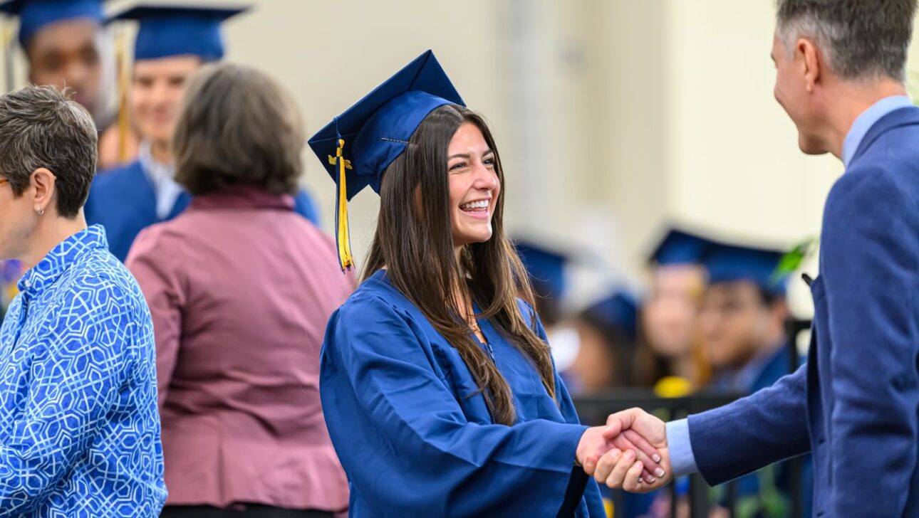 A graduate in a blue cap and gown shakes hands with a person in a blue suit during a graduation ceremony.