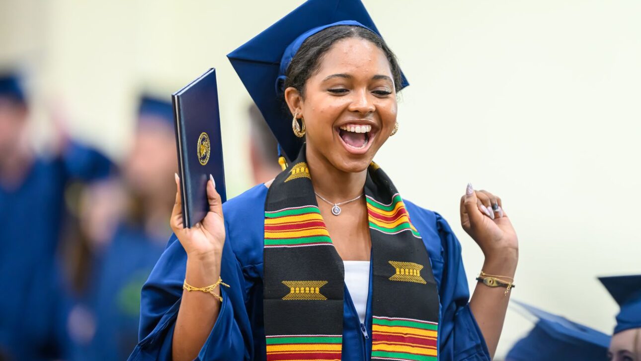 A graduate in a blue cap and gown celebrates, holding her diploma and wearing a colorful stole.