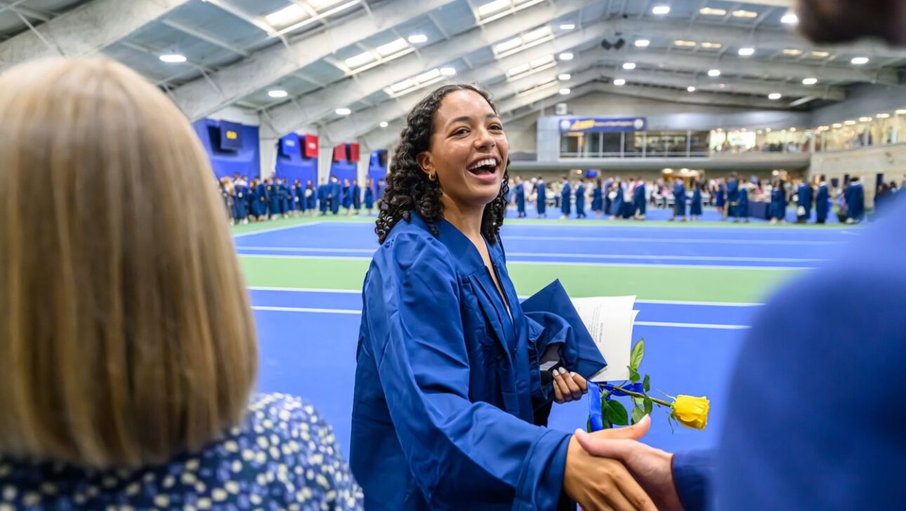 Graduate in blue gown smiles and shakes hands while holding a yellow rose and diploma. Indoor ceremony with other graduates and attendees in the background.