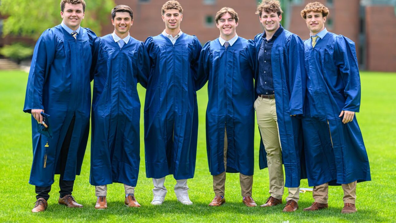 Six young men standing on grass wearing blue graduation gowns and smiling at the camera.