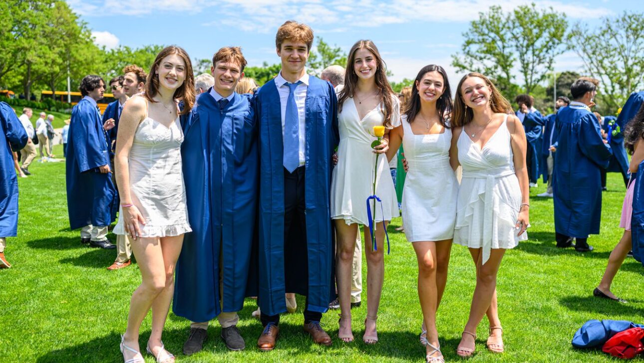 A group of young adults, four in white dresses and two in graduation gowns, stand together on a grassy field during a sunny day, with other people and trees in the background.