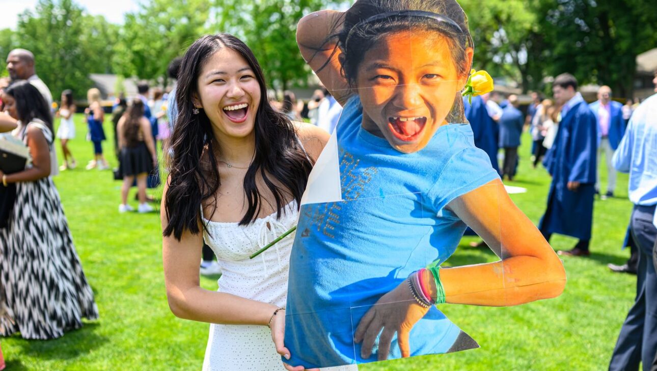 A young woman holding a large cutout of a child making a playful pose, with people gathered in the background on a sunny day.
