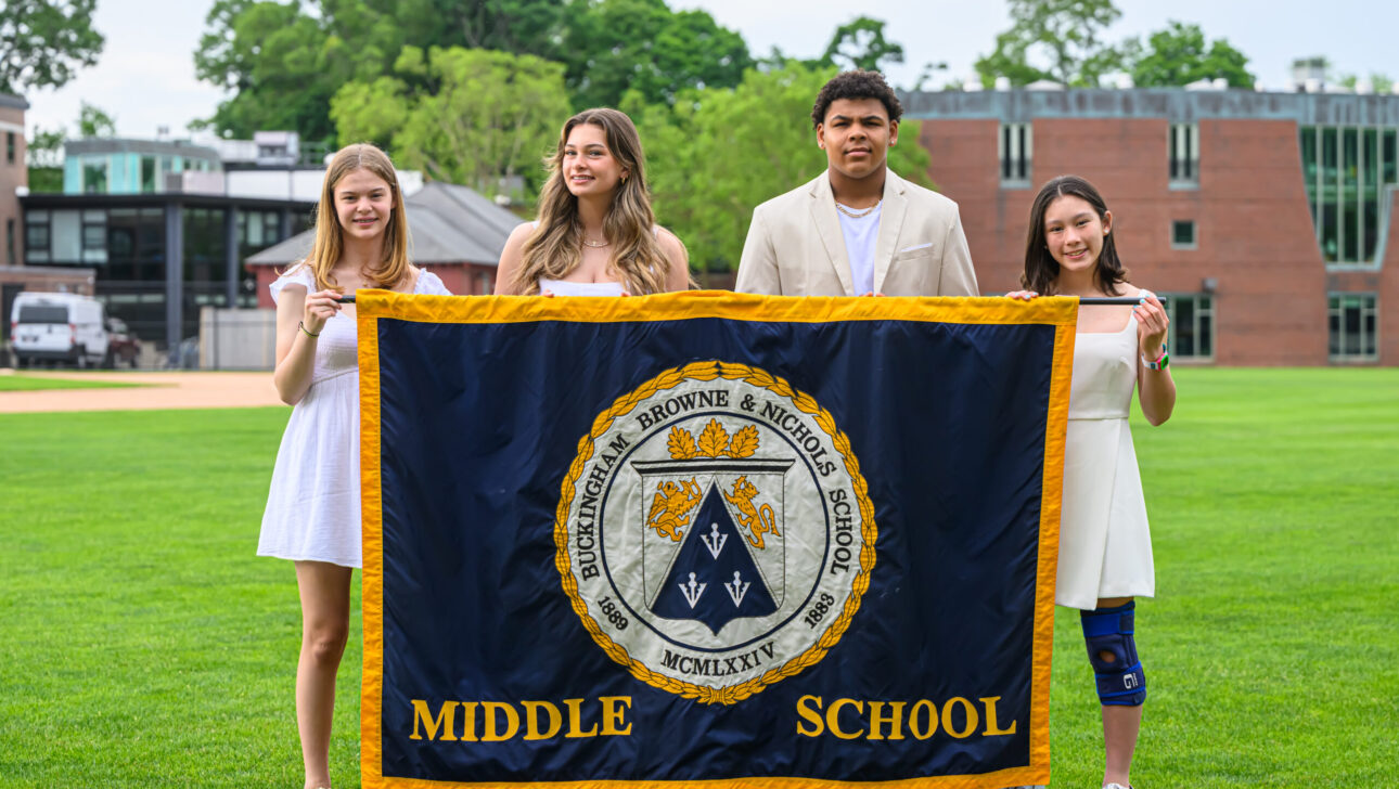 Four middle school students, three girls in white dresses and one boy in a beige suit and tie, hold a large blue and yellow school banner on a grassy field with buildings in the background.