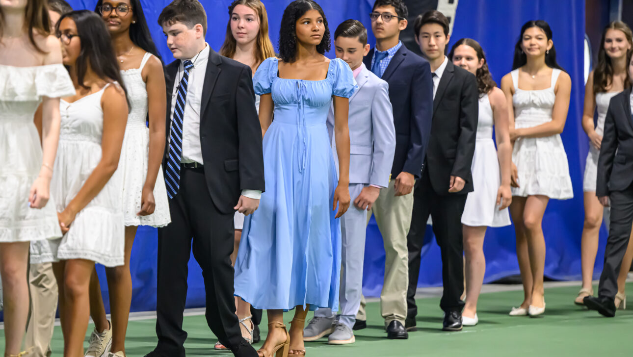 A group of young adults dressed in semi-formal attire, with men in suits and women in dresses, walk in a single-file line indoors.