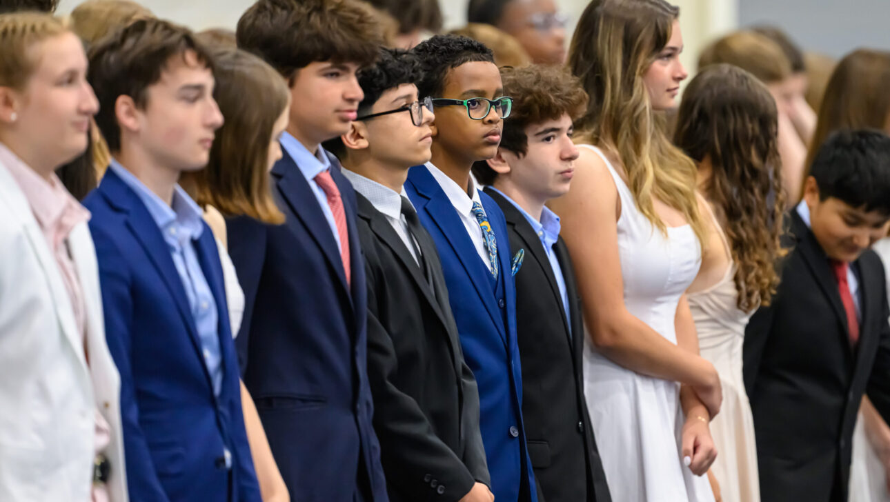 A group of young people is standing in a row, with some wearing formal suits and others in dresses, during what appears to be a formal event or ceremony.