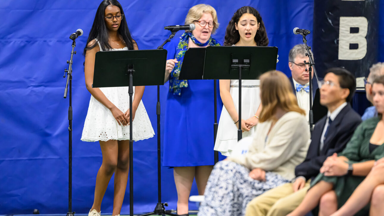 Three people stand at music stands with microphones, singing. Two women and one girl are seated in the audience in the foreground.