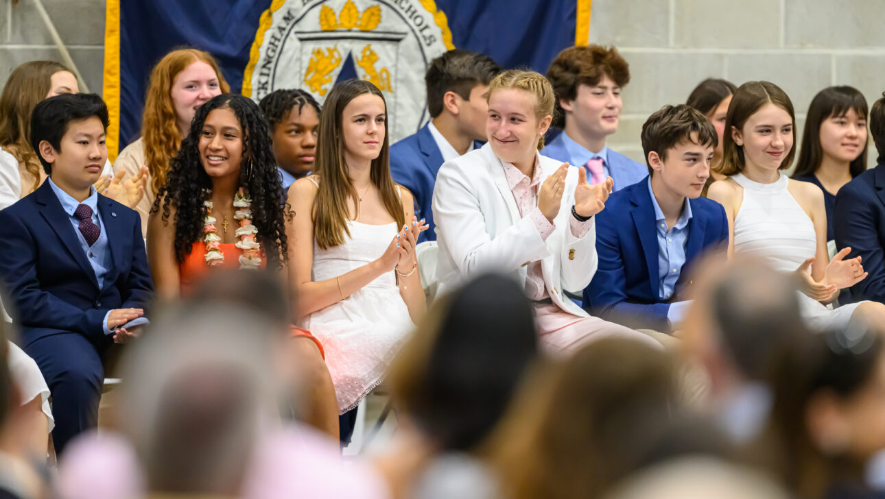 A group of students dressed formally sit in chairs at an event, some clapping.