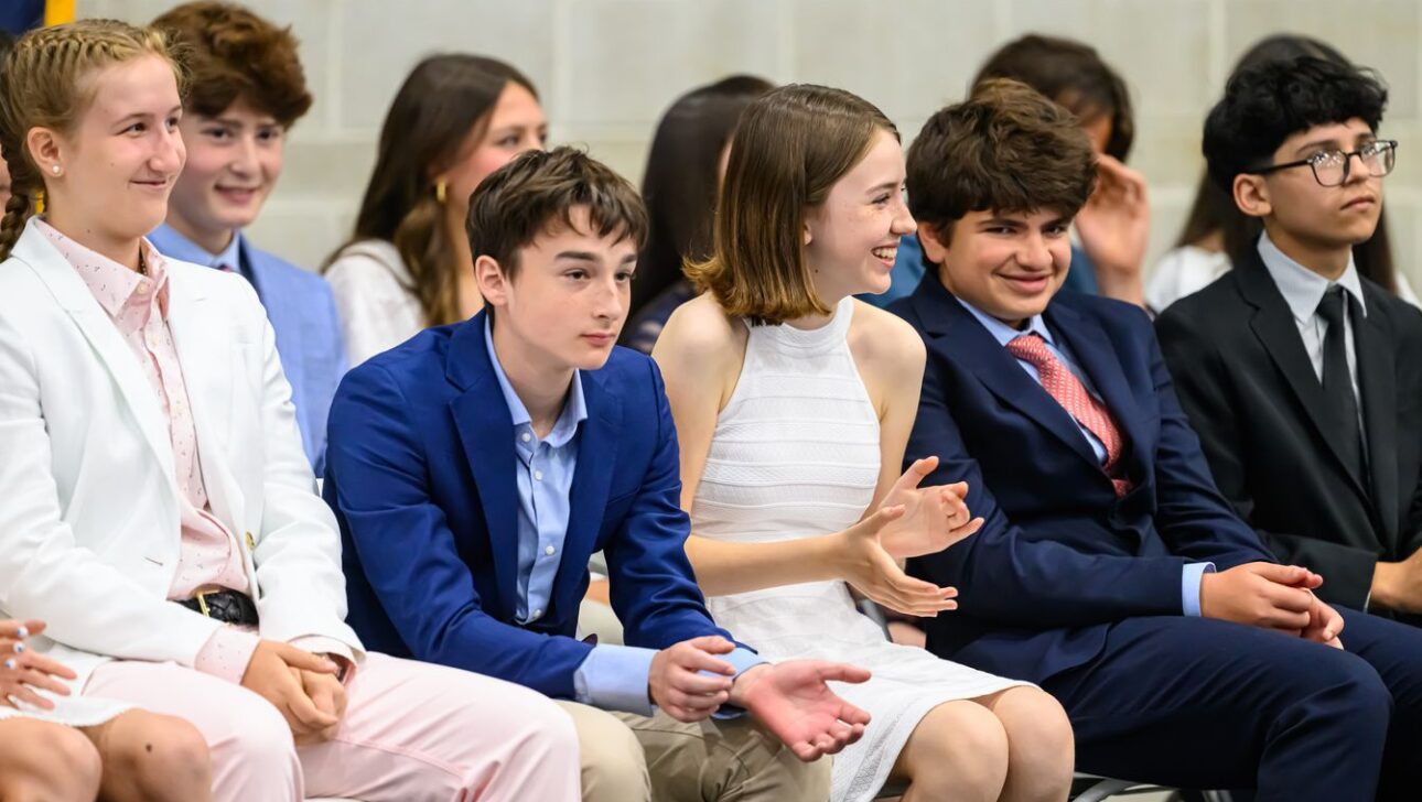 A group of young people sit in an audience, dressed in formal attire. Some are smiling and clapping, suggesting they are watching a performance or attending an event.
