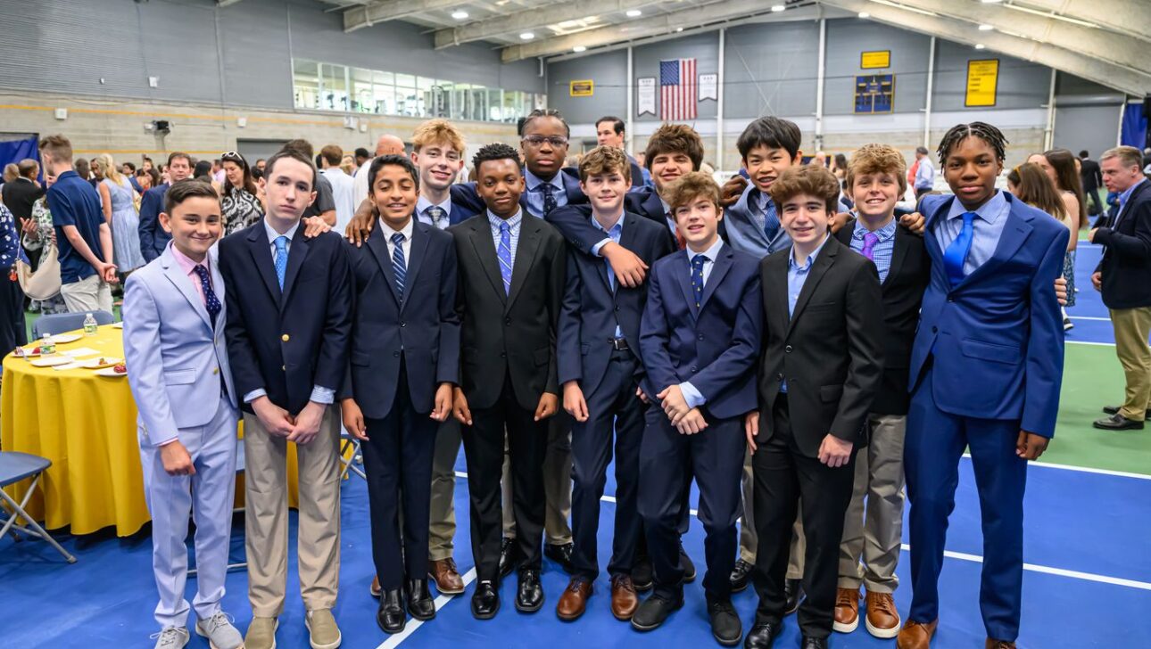 A group of young boys in formal attire standing together and posing for a photo in an indoor setting with other people in the background.