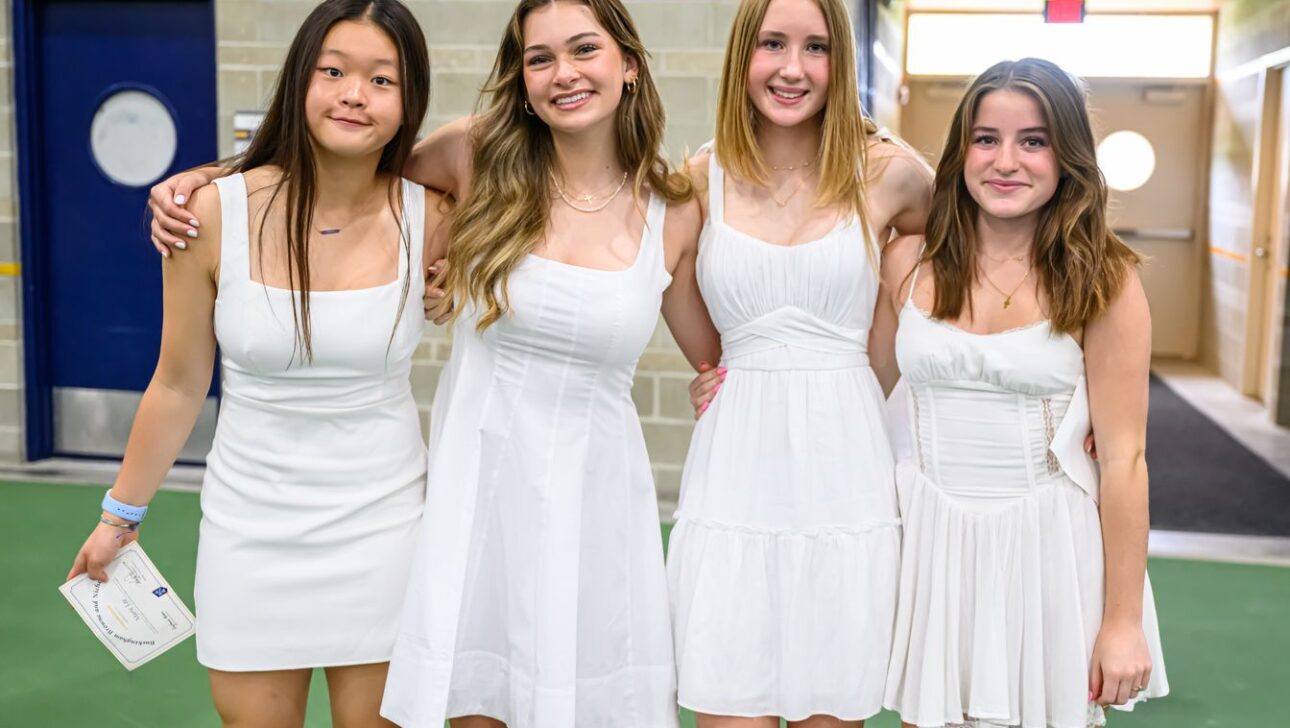 Four young women wearing white dresses stand side by side, smiling at the camera in an indoor setting with a blue doorway in the background.