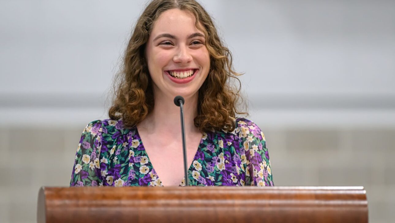 A person with curly hair and a floral dress smiles while standing at a podium with a microphone.