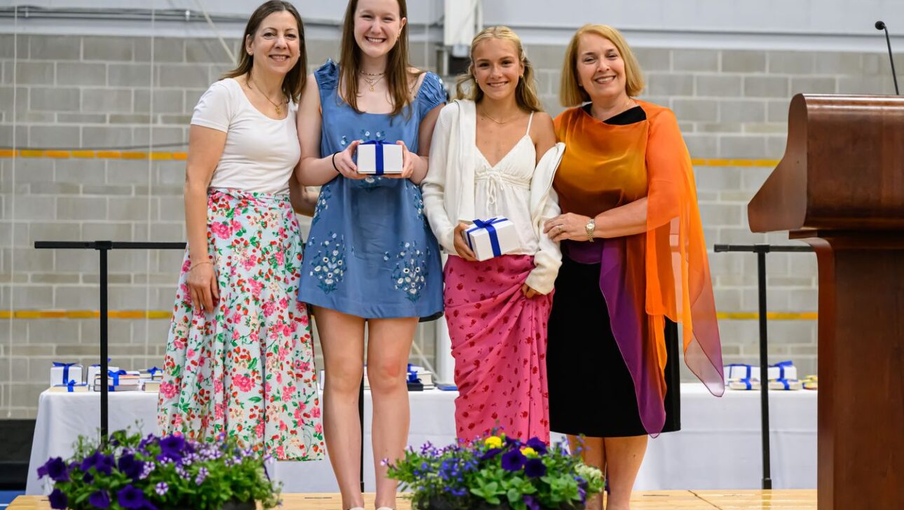 Four women stand on a stage, two of whom are holding gift boxes with ribbons.