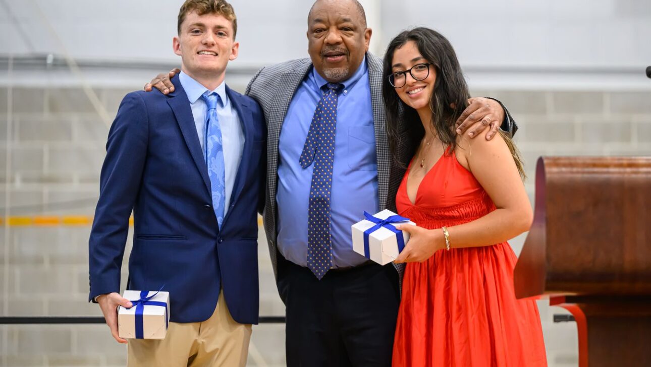 Three people pose for a photo at an event; the person in the center has an arm around each of the other two individuals, who are holding gift boxes.
