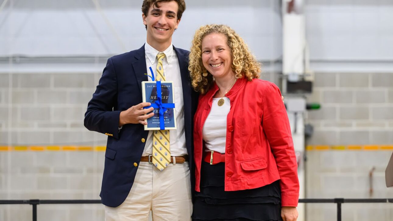 A young man in a navy blazer holding a blue ribbon award stands next to a smiling woman in a red jacket in a brightly lit indoor space with a gray block wall background.