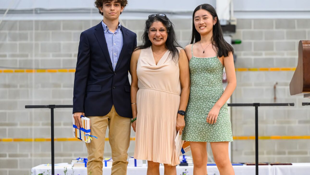 Three people stand together in a gymnasium: a young man in a blazer holding a book, a woman in a beige dress, and a young woman in a green dress.