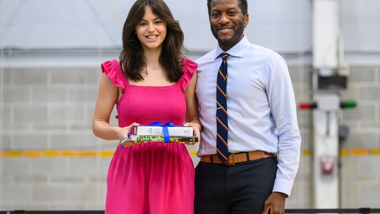 Two people stand side by side indoors, smiling. One is holding a set of books tied with a ribbon.