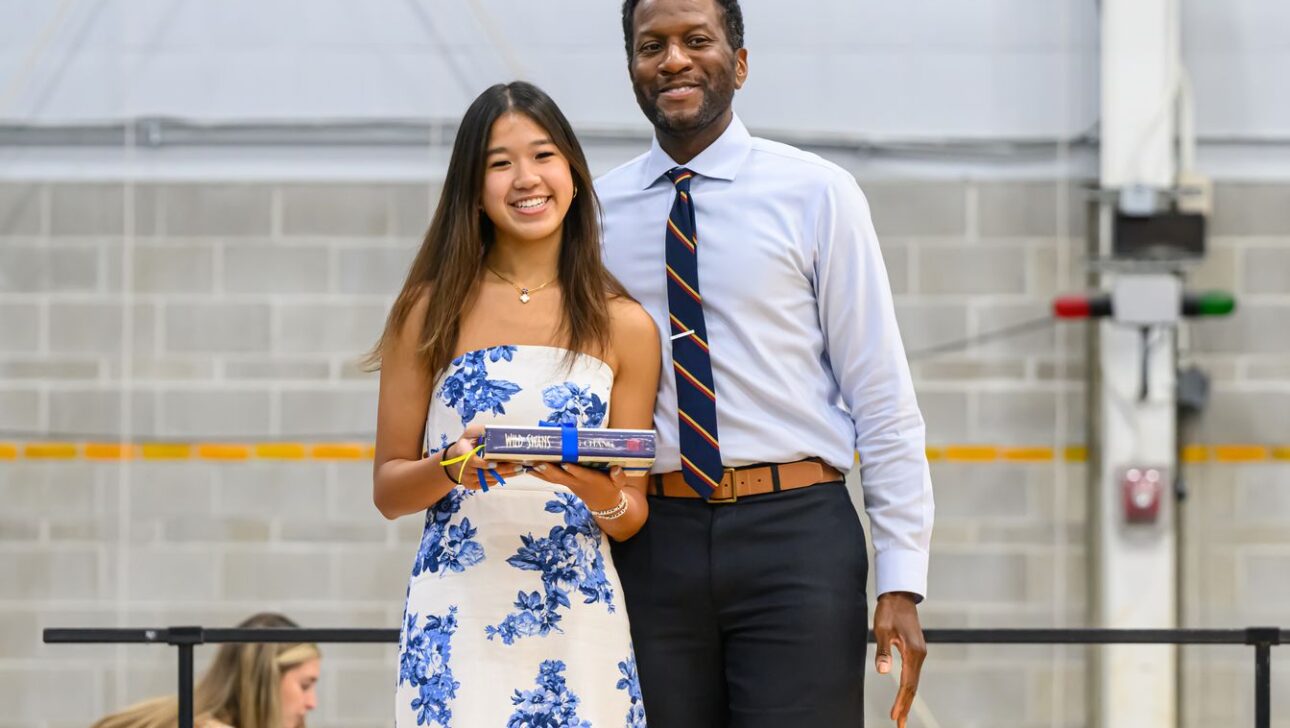 A young woman in a blue and white dress holds a boxed item and stands next to a smiling man in a tie, both posing for a photo indoors.