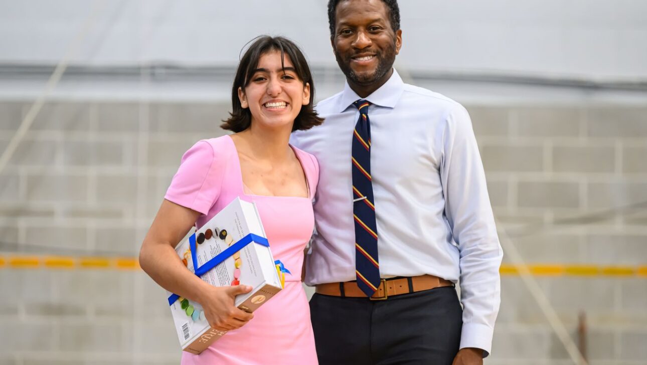 Two people stand close together, smiling at the camera. The person on the left is holding a gift box with a blue ribbon.