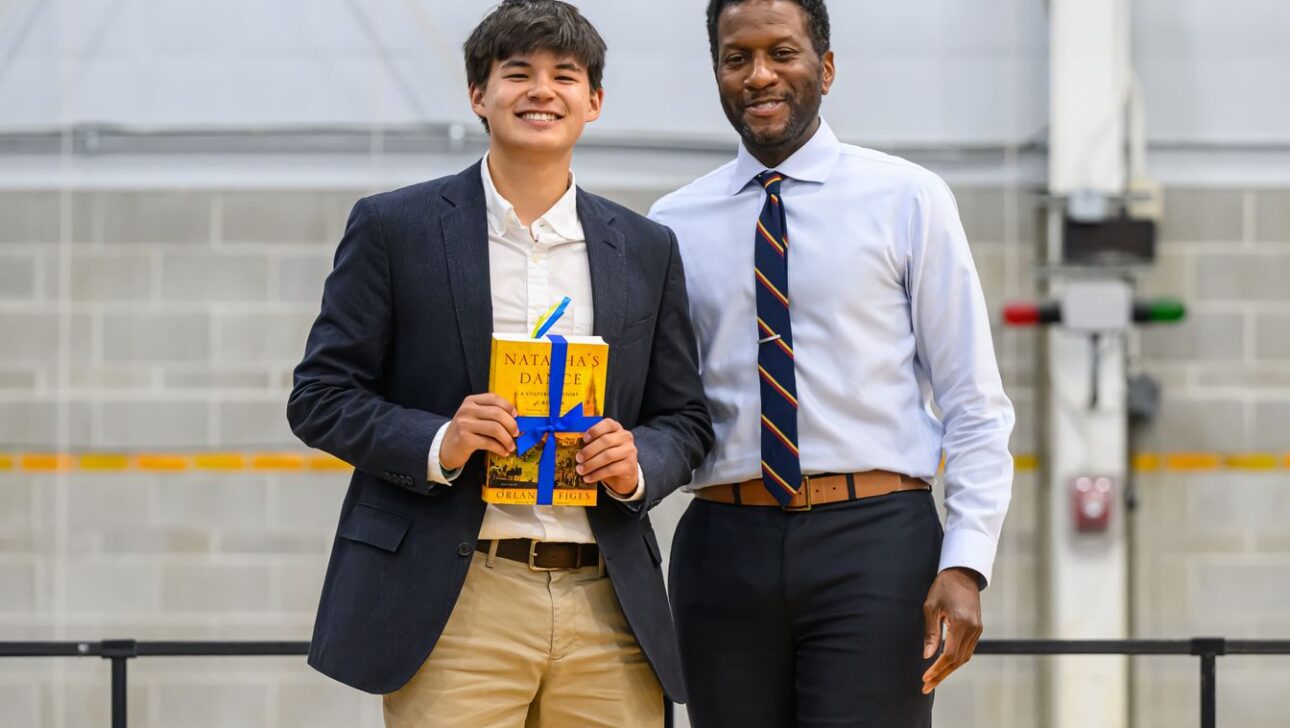 Two men are smiling while standing together indoors, one holding a yellow book.