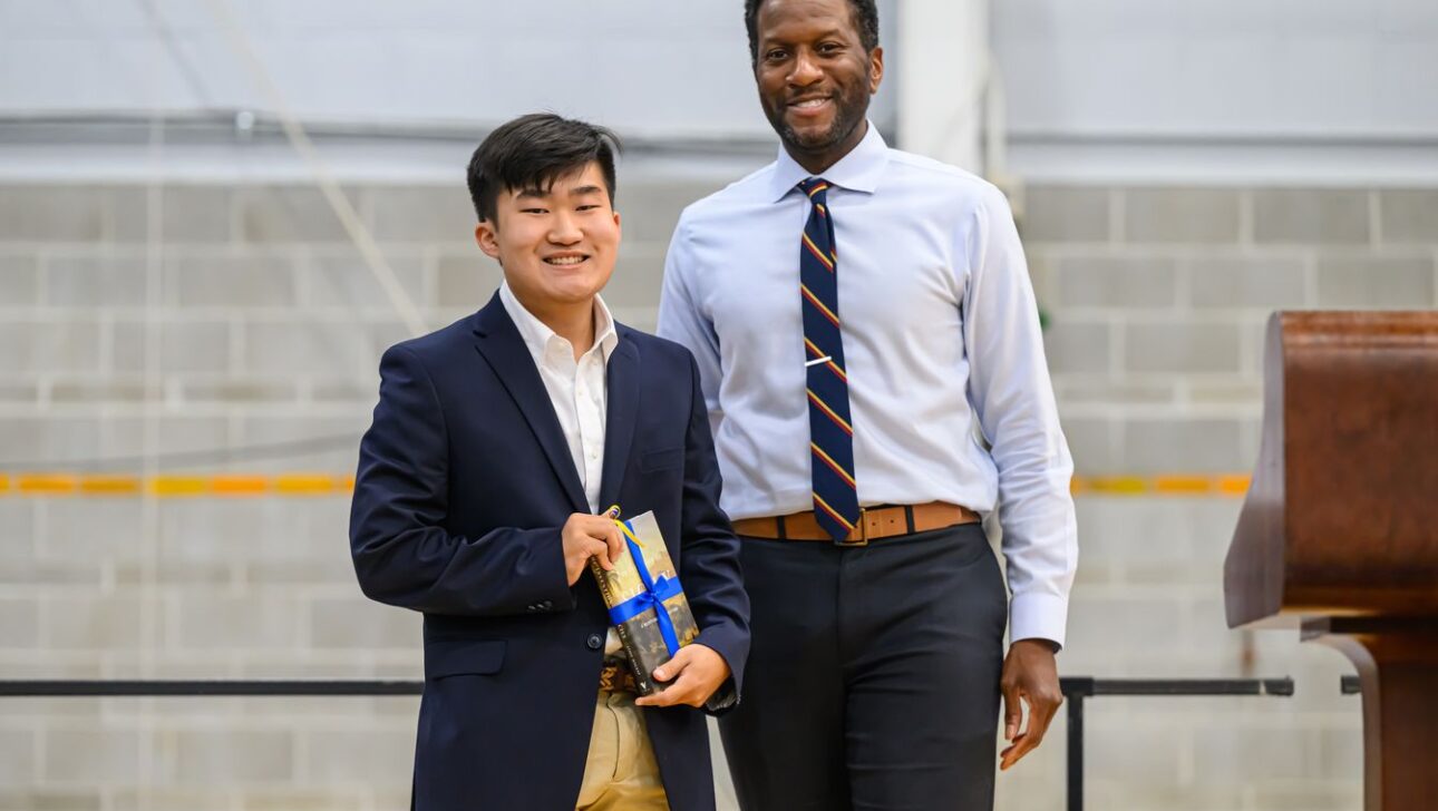 Two individuals stand together, one holding a gift. Both are dressed in business attire, with a background of a brick wall and a podium.