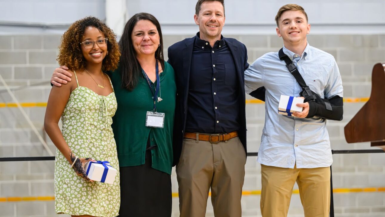 Four people stand side by side, smiling at an indoor event. Two of them hold gift boxes, and one has their arm in a sling.