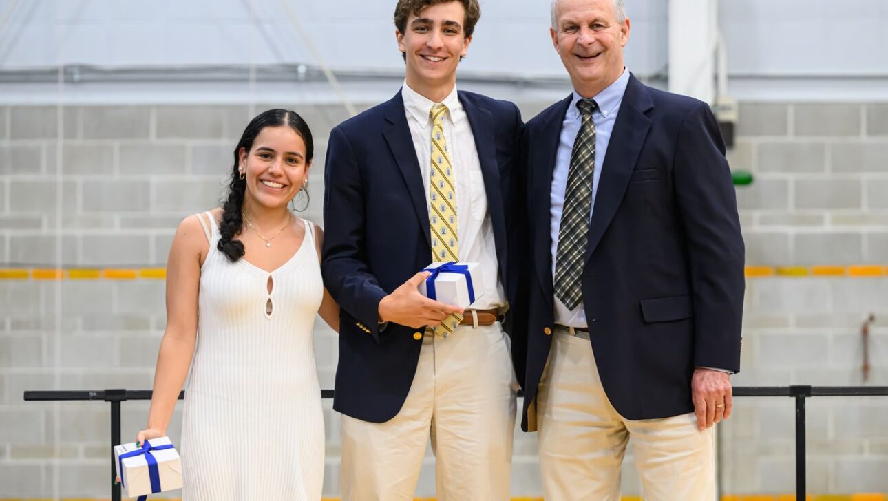 Three people in formal attire smiling and holding gift boxes, posed in a gymnasium.