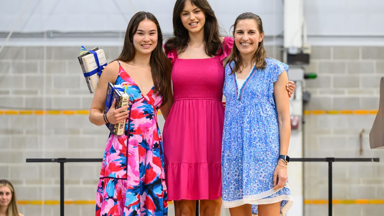 Three women standing together, all smiling, in colorful dresses; one woman holds a stack of books and a wrapped gift.