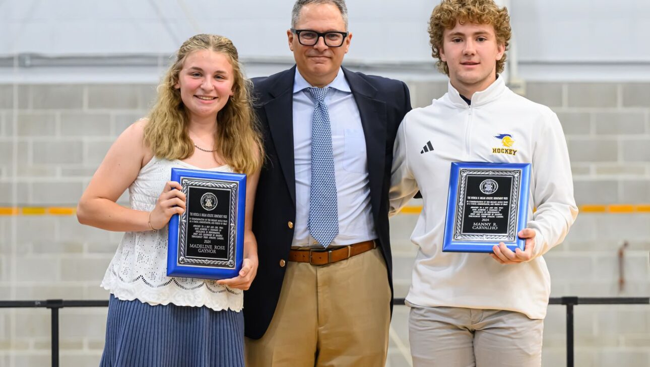 Three people pose for a photo; two young individuals, one holding a tennis plaque and the other a hockey plaque, stand on either side of an older person wearing a suit.