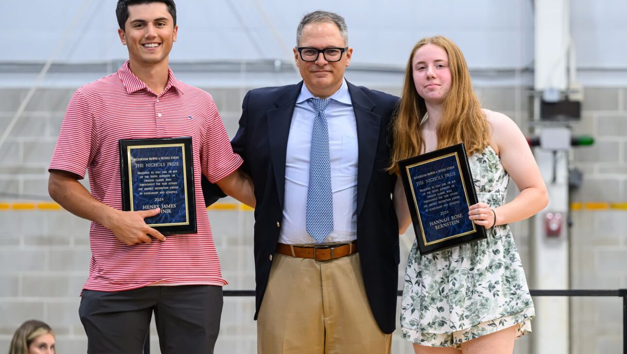 Three individuals stand side by side; the man in the center wears a suit, and the two on either side hold plaques.