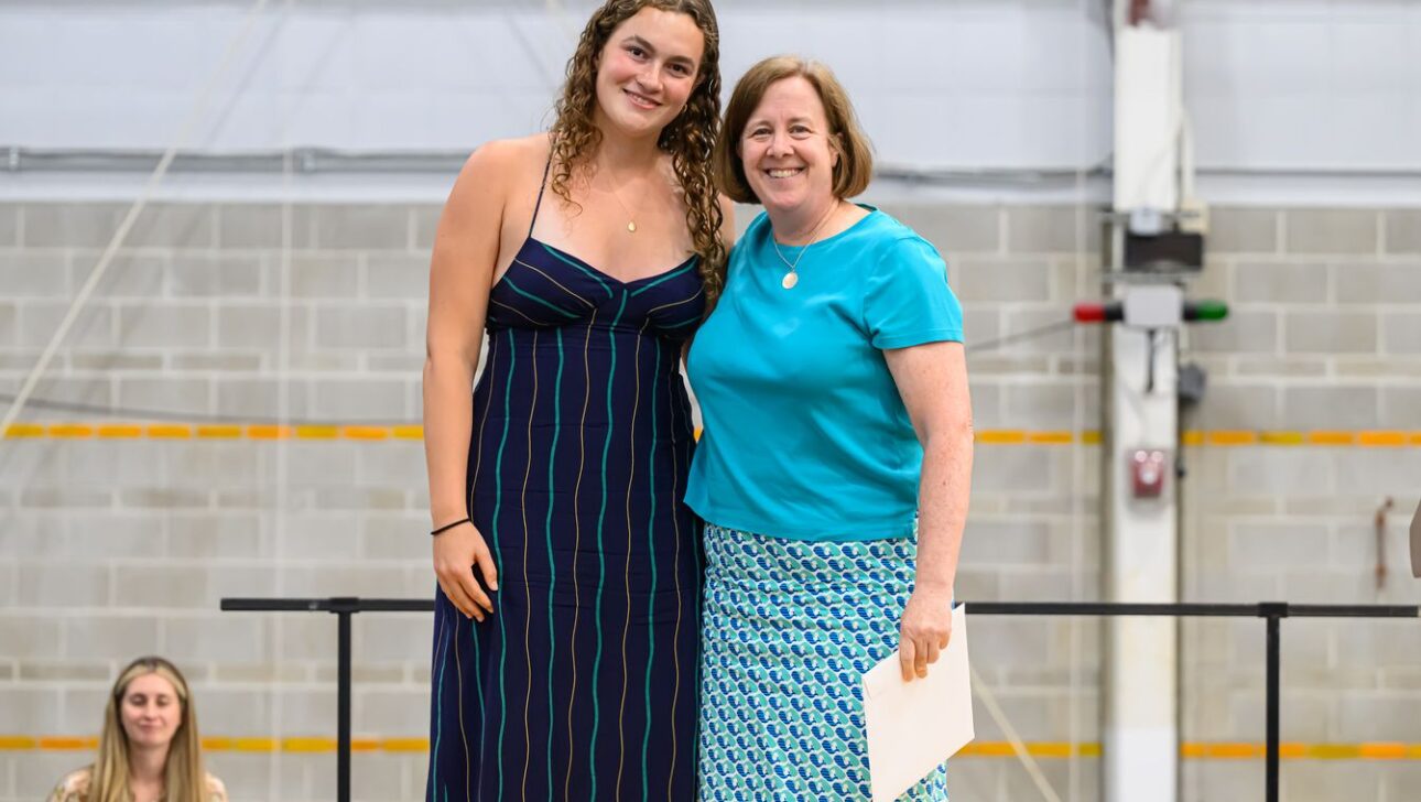 Two women smiling for a photo indoors; one wears a dark striped dress, the other a blue top and patterned skirt, while holding a certificate in their right hand.