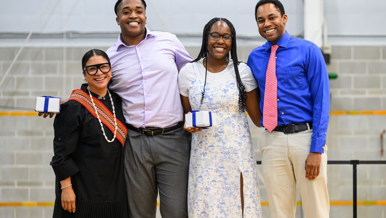 Four people are smiling and standing side by side. Two individuals in the center are each holding a small gift box.