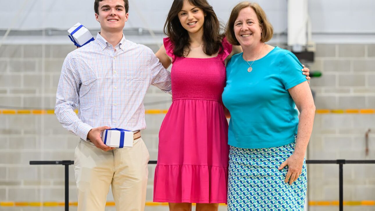 Three people stand indoors. One holds a gift box.