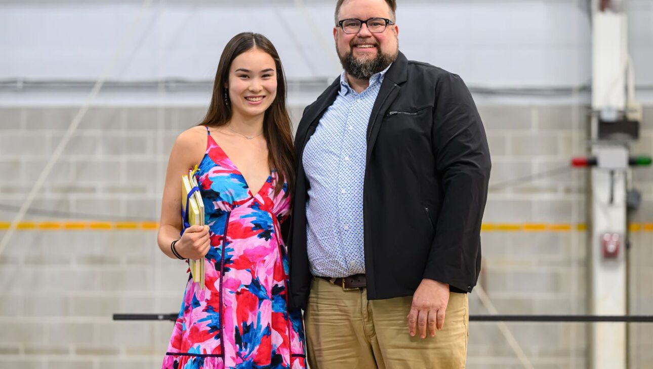 A woman in a colorful floral dress stands next to a man in a black jacket and light blue shirt, both smiling, with a gray industrial background.