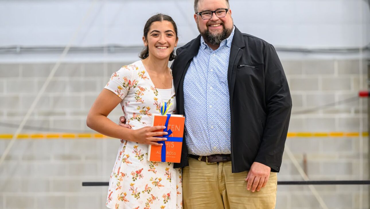 Two people smiling, one holding a book and wearing a floral dress, the other in a blue shirt, black jacket, and beige pants, standing in an indoor space with a concrete wall and yellow stripe.