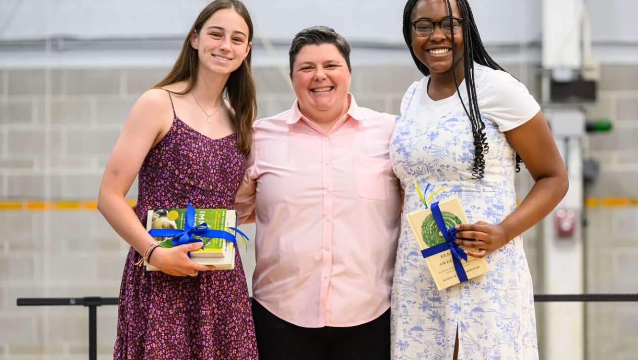 Three individuals stand together indoors, smiling, with two holding gifts wrapped with blue ribbons.