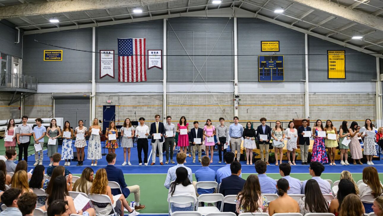 A group of people stand in a row holding certificates in an indoor gymnasium, with seated individuals watching them.