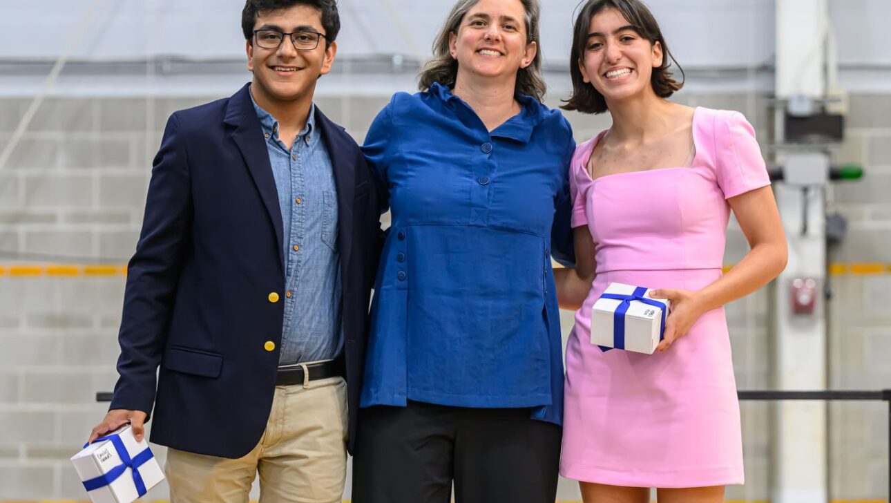 Three individuals stand indoors, smiling. Two hold identically wrapped gifts. The person in the center has their arms around the other two.