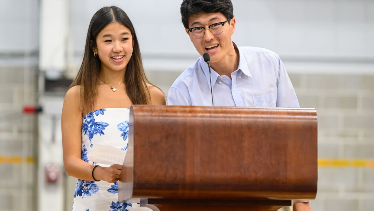 Two individuals stand behind a wooden podium, speaking into a microphone.