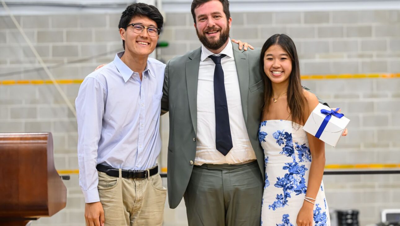 Three individuals posing together, with the person in the middle wearing a suit and the other two dressed in casual attire; one holding a gift box.