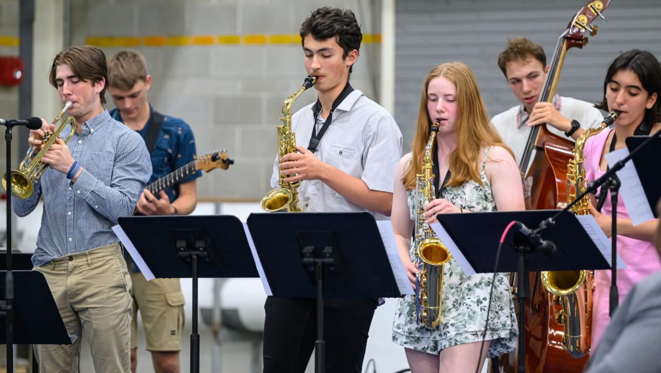 A group of young musicians performing with various instruments including saxophones, a trumpet, guitar, and double bass in a rehearsal setting.