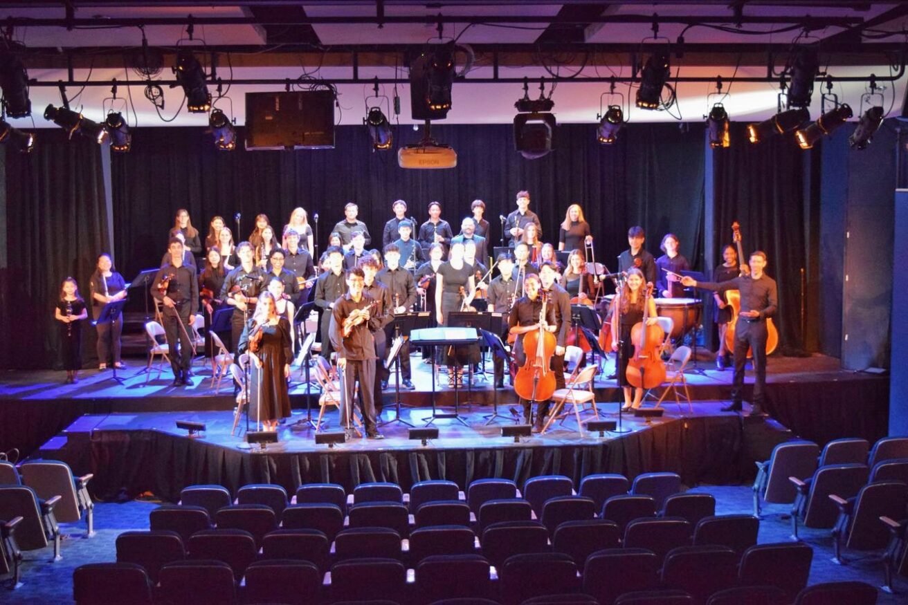 A full orchestra performs on stage with various string, woodwind, brass, and percussion instruments. Choristers stand behind the instrumentalists.