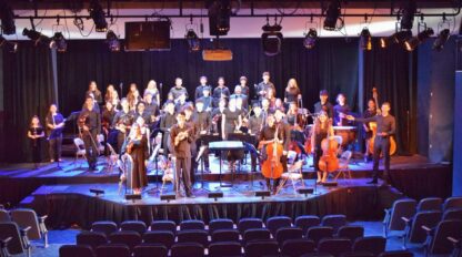 A full orchestra performs on stage with various string, woodwind, brass, and percussion instruments. Choristers stand behind the instrumentalists.