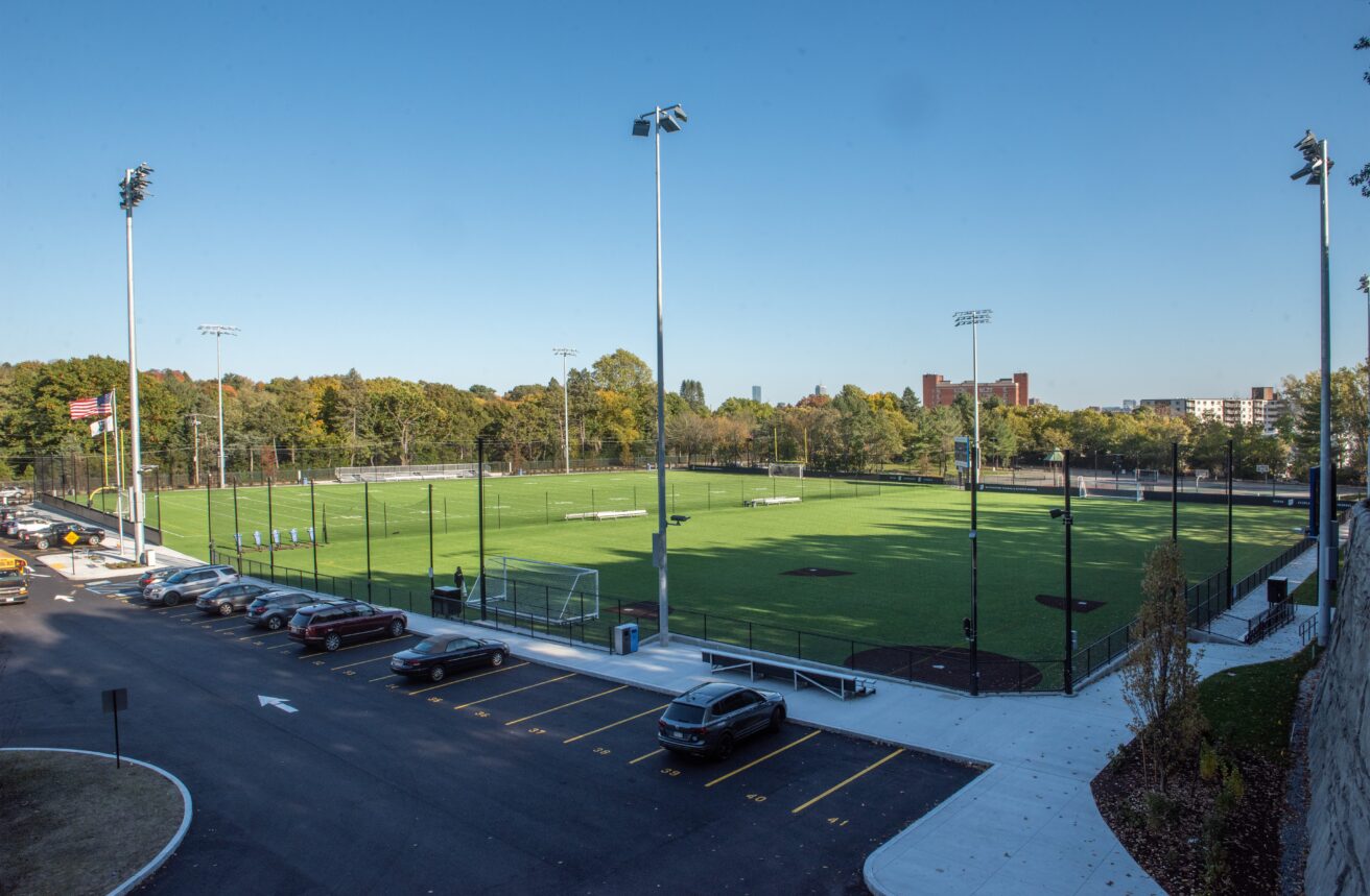 A green sports field with surrounding lights, adjacent parking lot, and trees in the background under a clear blue sky.
