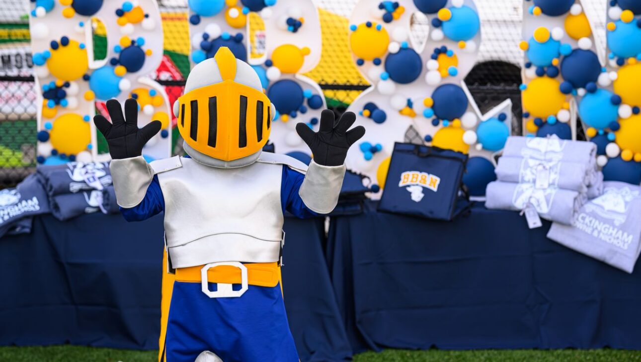 A mascot in a knight costume poses with raised hands on a grassy field. Behind are decorated letters and tables with folded clothing items.