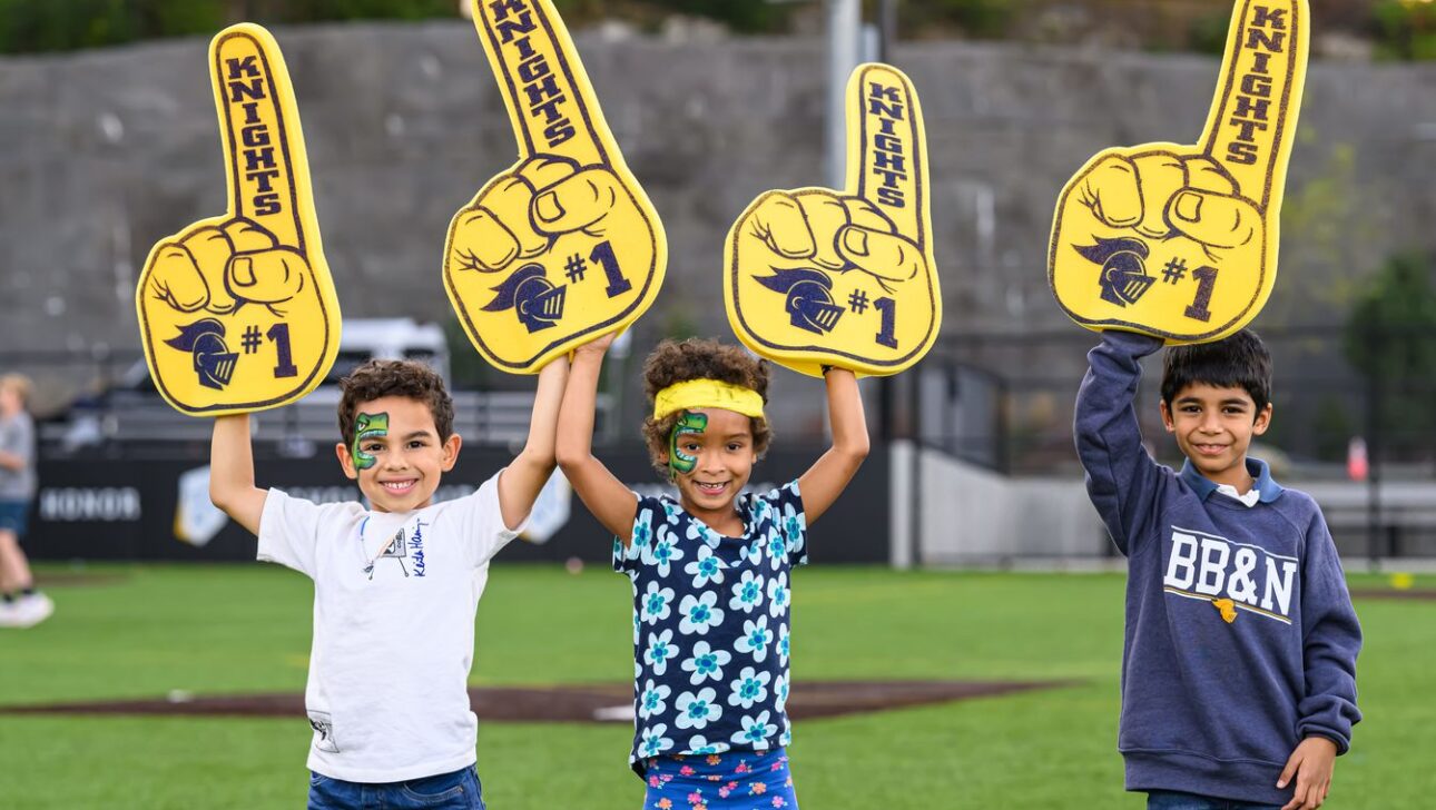 Three children stand on a sports field holding large foam fingers with "Knights #1" written on them. They have face paint and are smiling.